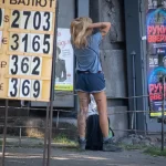Фотография девушки с татуировкой на ноге, стоящей у входа в здание. Татуировка изображает цветы и надпись. Девушка одета в шорты и футболку.
