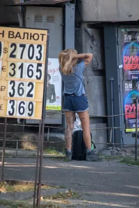 Фотография девушки с татуировкой на ноге, стоящей у входа в здание. Татуировка изображает цветы и надпись. Девушка одета в шорты и футболку.
