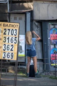 Фотография девушки с татуировкой на ноге, стоящей у входа в здание. Татуировка изображает цветы и надпись. Девушка одета в шорты и футболку.