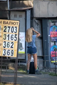 Фотография девушки с татуировкой на ноге, стоящей у входа в здание. Татуировка изображает цветы и надпись. Девушка одета в шорты и футболку.