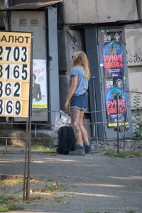Фотография девушки с татуировкой на ноге, стоящей на улице возле здания. Девушка в шортах и футболке, с рюкзаком.