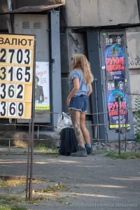 Фотография девушки с татуировкой на ноге, стоящей на улице возле здания. Девушка в шортах и футболке, с рюкзаком.