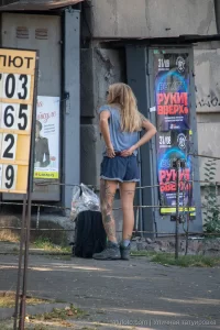 Фотография девушки с татуировкой на ноге, стоящей на улице возле здания. Девушка в шортах и футболке, с рюкзаком.