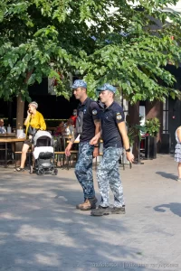 Фотография двух мужчин в военной форме с видимыми татуировками на руках, идущих по улице. На заднем плане деревья и люди.