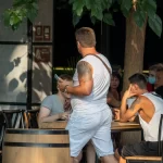 Фотография двух мужчин, один из которых с татуировкой на плече, сидящих за столиком в уличном кафе. Татуировка выполнена в темных тонах.