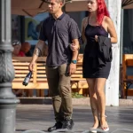 Фотография мужчины с татуировкой на предплечье и женщины с красными волосами, идущих вместе. Мужчина держит в руке что-то.