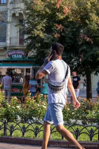 Фото тату на руке мужчины, выполненное в стиле графика. Видны детализированные изображения и темные контуры. Мужчина стоит возле кофейного трама на улице.