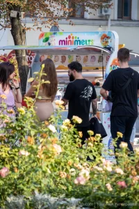 Фото мужчины с татуировками на руках и плечах, стоящего возле киоска с мороженым. Видны детализированные рисунки татуировок в темных тонах, включая череп.