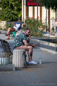 Фото тату на руке мужчины, сидящего на бетонном блоке в парке, вид сбоку, летний день, городская среда.