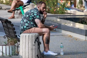 Фото тату на руке мужчины, сидящего на бетонном блоке в парке, вид сбоку, летний день, городская среда.
