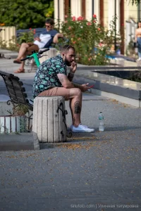 Фото тату на руке мужчины, сидящего на бетонном блоке в парке, вид сбоку, летний день, городская среда.