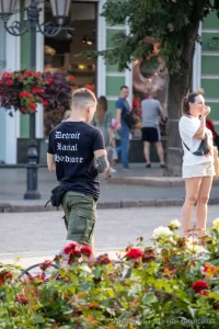 Фотография мужчины с татуировкой на руке, идущего по улице. Татуировка выполнена в темных тонах и имеет сложный узор. Снимок сделан в солнечный день.