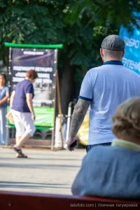 Фото мужчины с татуировкой на предплечье, стоящего на улице. Видна часть татуировки с розами и растительным орнаментом. Общий план, городской фон.