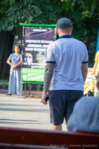 Фото мужчины в кепке с татуировкой на предплечье в стиле blackwork. Мужчина стоит спиной к камере, разговаривая с кем-то.