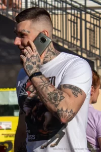 Фото мужчины с татуировками на руках и шее, разговаривающего по телефону. Видны детализированные рисунки, включая черепа и цветы.