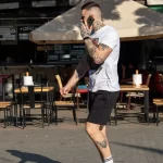 Фотография мужчины с татуировками на руках и ногах, разговаривающего по телефону на фоне кафе. Демонстрация современного стиля татуировки.