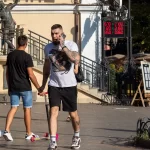 Фото мужчины с татуировками на руках и шее, едущего на самокате на улице. Видны детализированные рисунки, включая всадника на коне.