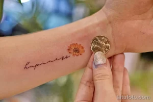 Минималистичная татуировка цветка и надписи 'Happiness' на запястье, выполненная в нежных тонах. Рисунок выглядит очень романтично и позитивно.