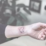 Небольшая, минималистичная татуировка котенка на запястье, выполненная тонкими черными линиями. Рядом с котенком изображено маленькое сердечко.