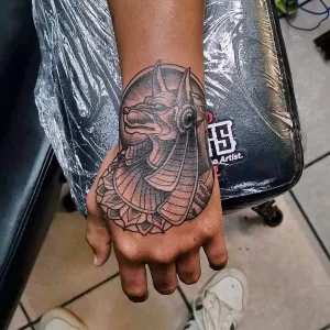 Фото тату на руке: детальное изображение головы бога Анубиса в египетском стиле. Черно-серый рисунок татуировки с четкими линиями и проработкой деталей.