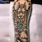 Фото тату Анубиса на руке, выполненное в ярких цветах с использованием синего, золотого и черного. Изображение включает в себя символы Уаджет и весы правосудия.