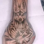Чёрно-серая татуировка Анубиса на руке, выполненная в реалистичном стиле. Детализированный рисунок татуировки с изображением бога.
