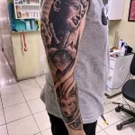 Фото тату, сочетающее изображение Анубиса и фараона в черно-сером стиле на руке, с детализированными лицами и элементами древнеегипетской культуры.