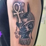 Фото татуировки Анубиса с весами и символом Анх на плече. Изображение выполнено в черно-серых тонах с высокой детализацией.