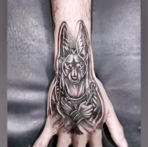 Черно-серое фото татуировки Анубиса на руке мужчины. Детализированный рисунок изображает голову шакала с атрибутами бога.