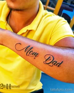 Черно-белое фото татуировки на руке с надписью "Mom Dad", выполненной красивым шрифтом. Простая и трогательная татуировка, посвященная родителям.