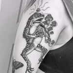 Черно-белое фото татуировки на плече с изображением лягушки, сидящей на грибах. Рядом с лягушкой изображена бабочка. Детализированный рисунок в сюрреалистичном стиле.