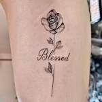 Фото тату на руке: реалистичная роза с зелеными листьями и надписью 'Blessed' каллиграфическим шрифтом.