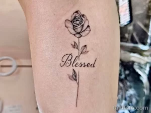 Фото тату на руке: реалистичная роза с зелеными листьями и надписью 'Blessed' каллиграфическим шрифтом.