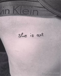 Черно-белое фото минималистичной татуировки с надписью 'She is art' на боку. Изящный шрифт и лаконичный дизайн.