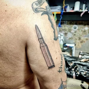 Фото минималистичной татуировки в виде пули, выполненной в технике blackwork на предплечье, с добавлением надписи 'thank'.