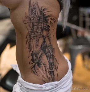Фото тату Анубиса на боку женщины. Детализированный рисунок в черно-серых тонах, изображающий египетского бога с мечом и украшениями.