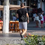 Фото тату на руке мужчины, идущего по улице. Видна часть татуировки в темных тонах, с растительными элементами.