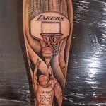 Фото тату на руке: детализированный портрет Майкла Джордана с логотипом Lakers и видом на город. Выполнено в черно-серых тонах.