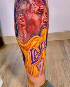 Фото цветной татуировки с портретом Коби Брайанта в форме Los Angeles Lakers на ноге, с использованием ярких и насыщенных цветов.