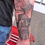 Черно-серая татуировка на руке с изображением Майкла Джордана в прыжке, окруженного надписями и баскетбольной сеткой. Динамичный рисунок татуировки.