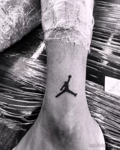 Фото тату на лодыжке с изображением логотипа Jumpman (силуэт Майкла Джордана в прыжке). Черная татуировка в минималистичном стиле.