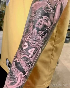 Фото тату портрета Коби Брайанта в форме Lakers на предплечье, с элементами розы и брызгами краски. Реалистичный рисунок татуировки.