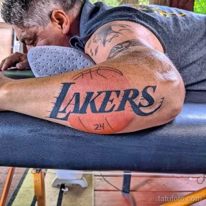 Фото тату на руке с изображением логотипа баскетбольной команды Lakers и номера 24, выполненное в черных тонах.