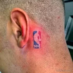 Небольшое цветное фото татуировки с логотипом NBA, расположенной за ухом на шее. Детализированный рисунок татуировки.