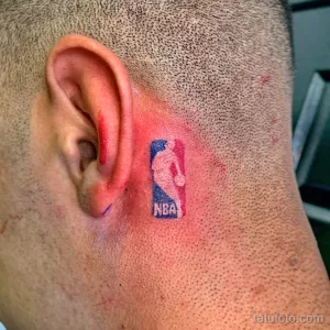 Небольшое цветное фото татуировки с логотипом NBA, расположенной за ухом на шее. Детализированный рисунок татуировки.