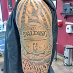 Фото тату с изображением баскетбольного мяча Spalding, логотипа Филадельфии 76ers и подписи, выполненное в черно-серых тонах на плече.