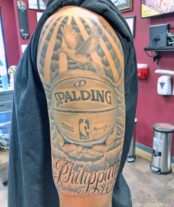 Фото тату с изображением баскетбольного мяча Spalding, логотипа Филадельфии 76ers и подписи, выполненное в черно-серых тонах на плече.