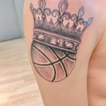Черно-серое фото тату на плече с изображением баскетбольного мяча, увенчанного короной, выполненное в реалистичном стиле.