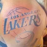 Фото татуировки на плече с изображением логотипа баскетбольной команды Los Angeles Lakers. Тату выполнено в черно-синих тонах.
