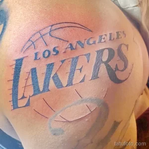 Фото татуировки на плече с изображением логотипа баскетбольной команды Los Angeles Lakers. Тату выполнено в черно-синих тонах.